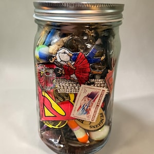 May include: A clear glass mason jar with a silver metal lid, filled with a collection of assorted small items including a red and blue embroidered patch, a postage stamp labeled Republique Rwandaise, a yellow graphic card, and a marble.