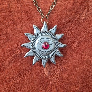 May include: Silver-toned sunburst pendant necklace with a red gemstone center. The pendant has a detailed design with pointed rays and a decorative circular center. The chain is a bronze color.
