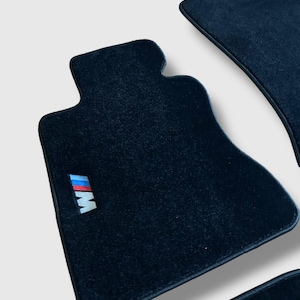 Könnte beinhalten: Dunkelblaue Fußmatten für Autos mit den Buchstaben "M" und "W" in Weiß und den Farben Blau, Rot und Hellblau. Die Matten bestehen aus einem weichen, strukturierten Material.