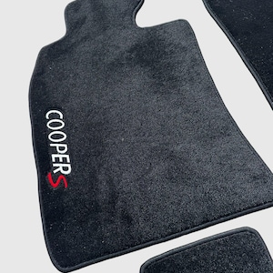 Op de afbeelding: Zwarte automatten met de tekst "COOPER S" geborduurd in wit en rood. De matten hebben een voorgevormde vorm en een zwarte rand. De afbeelding toont een set matten, waarschijnlijk voor een voertuig.