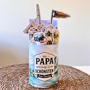 Puede incluir: Caja de regalo cilíndrica con temática de camping. La caja presenta ilustraciones de una cámara, una linterna, una tienda de campaña y una caña de pescar. El texto de la caja dice "Mit PAPA unterwegs ist am SCHÖNSTEN Zum Vatertag."