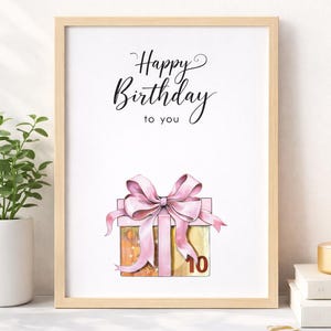 Puede incluir: Una impresión en acuarela enmarcada con las palabras "Happy Birthday to you" en una escritura elegante. La imagen presenta una caja de regalo con un lazo rosa y el número 10. La impresión está en un marco de madera clara, con una planta y una vela en el fondo.