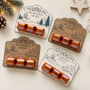 Könnte beinhalten: Vier dekorative Pappkarten mit einer Strichzeichnung von einem Schloss, Bäumen und Haselnüssen sowie dem Text "DREI HASELNÜSSE FÜR DICH". Jede Karte hält drei orange verpackte Ferrero Küsschen Schokoladen am unteren Rand.