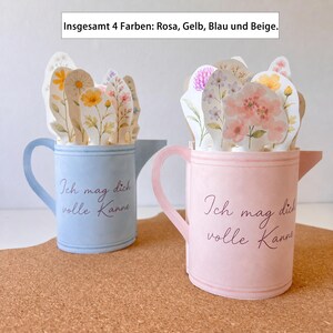 May include: Two paper watering cans, one light blue and one pink, each with a floral arrangement inside. The text "Ich mag dich volle Kanne" is written on the front of each can. The image also includes the text "Insgesamt 4 Farben: Rosa, Gelb, Blau und Beige."