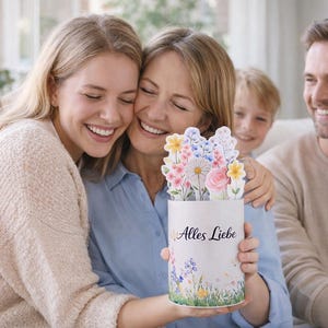 K&ouml;nnte beinhalten: Eine Familie umarmt sich und h&auml;lt eine wei&szlig;e Karte mit einem floralen Pop-up-Design. Die Karte tr&auml;gt den Text "Alles Liebe" und ist mit bunten Blumen und Gr&uuml;n geschm&uuml;ckt. Die Familie l&auml;chelt.