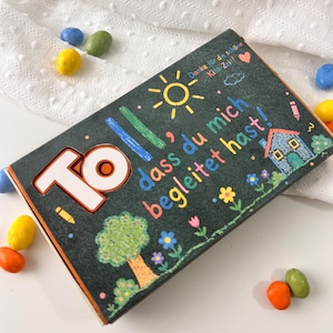 Peut inclure: Une boîte rectangulaire en bois avec un couvercle noir style ardoise orné de dessins colorés au crayon représentant un soleil, une maison, un arbre et des fleurs. Le texte indique "Toll, dass du mich begleitet hast!" et "Danke für die schöne Kita-Zeit!"