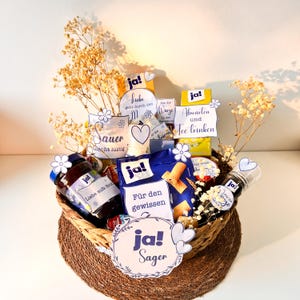 Könnte beinhalten: Ein Weidenkorb gefüllt mit verschiedenen Lebensmitteln und dekorativen Elementen. Der Korb enthält Gläser, verpackte Snacks und kleine Karten mit deutschem Text. Trockenblumen und Papierausstanzungen mit Blumen- und Herzmotiven ergänzen die Präsentation.