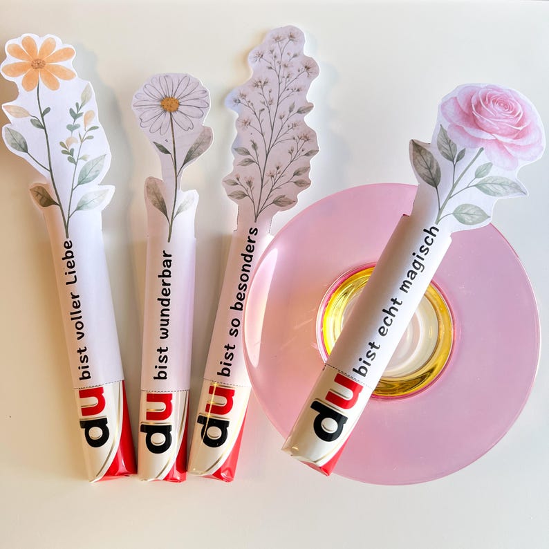 K&ouml;nnte beinhalten: Vier wei&szlig;e und rote S&uuml;&szlig;igkeitentuben mit floralen Aufs&auml;tzen und deutschem Text. Die Aufs&auml;tze zeigen eine G&auml;nsebl&uuml;mchen, eine Rose und andere florale Designs. Die S&uuml;&szlig;igkeitentuben sind auf einem rosa Teller angeordnet.