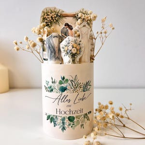 Könnte beinhalten: Eine zylindrische Geschenkbox aus Papier mit einer Aquarellillustration aus Eukalyptusblättern und dem Text Alles Liebe zur Hochzeit. Oben befinden sich ausgestanzte Papierfiguren eines Brautkleids, eines Anzugs und eines Blumenstraußes.