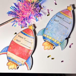 May include: Two rocket-shaped invitation cards, one red and one blue, with star accents and the text "Wann? Wo?" printed on them. The rockets have yellow flames and star-shaped tops. Confetti and a decorative wand are also visible.