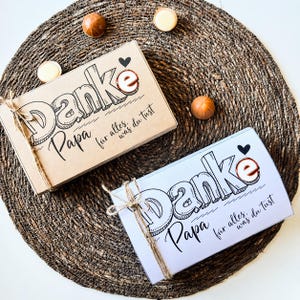 Peut inclure: Deux boîtes cadeaux avec le texte allemand "Danke Papa" (Merci Papa) et "für alles, was du tust" (pour tout ce que tu fais). Les boîtes sont attachées avec de la ficelle et décorées d'un cœur noir. Les boîtes sont sur un tapis tressé avec des chocolats.