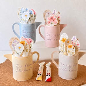 May include: A collection of pastel-colored watering cans, each holding floral arrangements. The cans are in shades of blue, pink, and beige, with handwritten text on them. Two small packets are in the foreground.