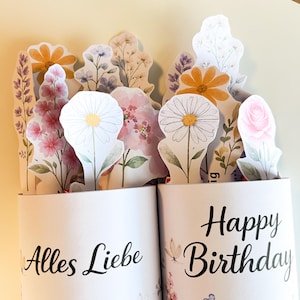 K&ouml;nnte beinhalten: Ein wei&szlig;er zylindrischer Beh&auml;lter mit den Aufschriften "Alles Liebe" und "Happy Birthday". Der Beh&auml;lter enth&auml;lt eine Sammlung von Papierblumen in verschiedenen Farben, darunter Rosa, Gelb und Wei&szlig;. Die Blumen sind so angeordnet, dass sie eine dekorative Darstellung bilden.