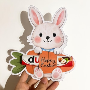 Puede incluir: Un conejito blanco de dibujos animados sosteniendo una zanahoria naranja con las palabras "Hoppy Easter". La zanahoria contiene una barra de chocolate. El conejito tiene mejillas y orejas rosas. La imagen es un recorte con fondo blanco.