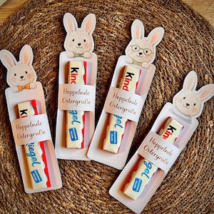 Könnte beinhalten: Vier Oster-Geschenkanhänger mit Hasen-Illustrationen, die Schokoriegel halten. Jeder Anhänger trägt die Aufschrift "Hoppelnde Ostergrüße" und "Kind" auf der Verpackung. Die Anhänger liegen auf einem geflochtenen Untergrund.
