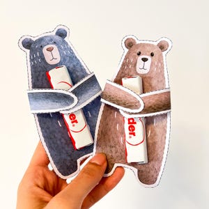Könnte beinhalten: Zwei papierförmige Bären-Geschenkehalter, einer blau und einer braun, halten jeweils einen weiß-roten Schokoriegel. Die Bären haben gestickte Umrisse und werden von einer Hand vor weißem Hintergrund gehalten. Auf dem Riegel ist das Wort "der" zu sehen.