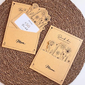 Pode incluir: Dois cartões de presente de papel kraft com recortes florais e texto manuscrito em preto. Um cartão diz "Mom" e tem um cartão de presente dentro. O outro cartão diz "Mama" e "Dank dir". Ambos os cartões têm detalhes de coração.