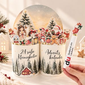 Puede incluir: Dos recipientes cilíndricos con una escena de bosque invernal y el texto en alemán 24 süße Kleinigkeiten Advents kalender. Las partes superiores están decoradas con recortes navideños que incluyen un ángel, Santa y un hombre de jengibre.