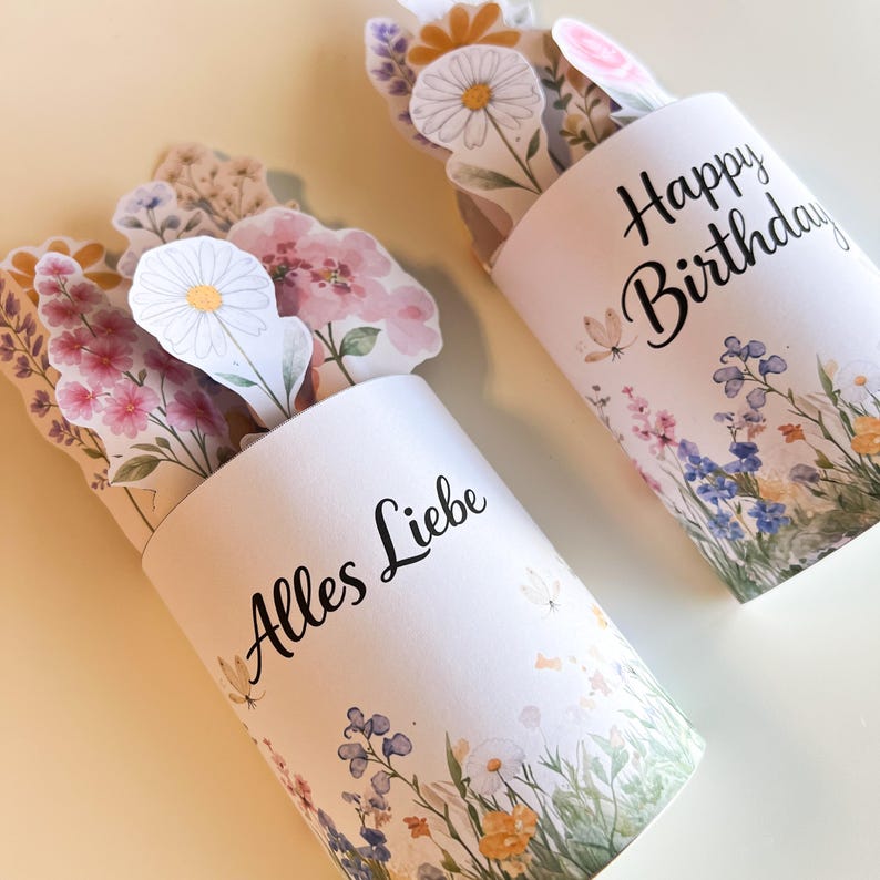 K&ouml;nnte beinhalten: Zwei wei&szlig;e zylindrische Beh&auml;lter mit floralen Designs und den Schriftz&uuml;gen "Alles Liebe" und "Happy Birthday" in schwarzer Schrift. Jeder Beh&auml;lter enth&auml;lt Papierblumen in verschiedenen Farben, darunter Rosa, Wei&szlig; und Gelb. Der Hintergrund ist hellgelb.
