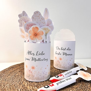 Könnte beinhalten: Zwei weiße zylindrische Behälter mit floralen Designs und Text. Einer sagt "Alles Liebe zum Muttertag" und der andere "Du bist die beste Mama". Florale Ausschnitte und zwei Süßigkeitenriegel mit floralen Akzenten sind ebenfalls vorhanden.
