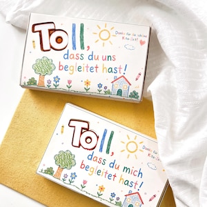 Pode incluir: Duas caixas retangulares de cartão branco com desenhos coloridos estilo giz de cera de uma árvore, flores, uma casa e um sol. O texto nas caixas diz "Toll, dass du uns begleitet hast!" e "Toll, dass du mich begleitet hast!" com um pequeno coração.