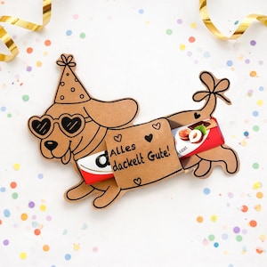 Könnte beinhalten: Ein brauner Geschenkhalter in Hundeform aus Pappe mit herzförmiger Sonnenbrille und Partyhut. Der Halter enthält zwei Schokoriegel. Der Text "Alles dackelt Gute!" ist auf dem Halter zusammen mit Herzillustrationen aufgedruckt.