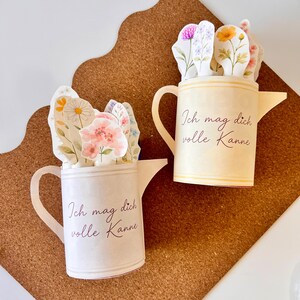 May include: Two paper watering cans, one cream and one off-white, each filled with floral cutouts. The cream can has yellow accents and the text "Ich mag dich volle Kanne". The off-white can has pink and white floral cutouts and the same text.