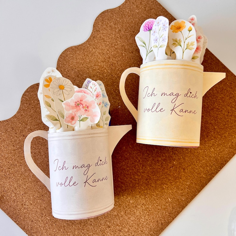 May include: Two paper watering cans, one cream and one off-white, each filled with floral cutouts. The cream can has yellow accents and the text "Ich mag dich volle Kanne". The off-white can has pink and white floral cutouts and the same text.