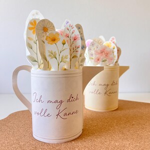 May include: Two paper flower arrangements in the shape of a mug and a watering can. The mug and watering can are off-white with the text "Ich mag dich volle Kanne" in burgundy. The flowers are watercolor-style with yellow, pink, and white blooms.