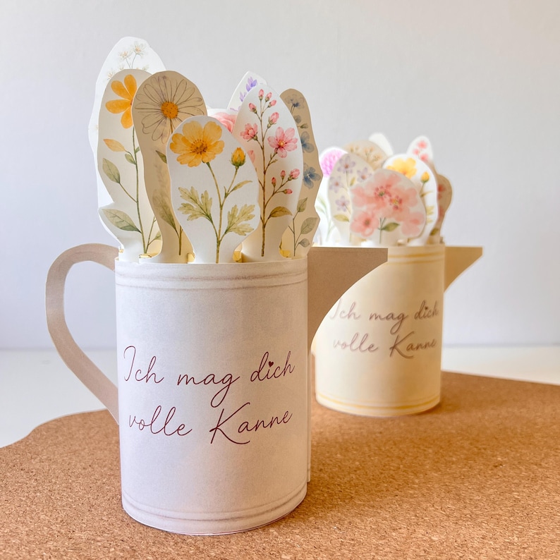 May include: Two paper flower arrangements in the shape of a mug and a watering can. The mug and watering can are off-white with the text "Ich mag dich volle Kanne" in burgundy. The flowers are watercolor-style with yellow, pink, and white blooms.