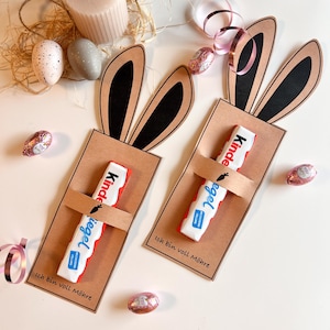 May include: Two Easter-themed gift card holders shaped like bunny ears, each holding a Kinder chocolate bar. The holders are brown with black ears and the text "Ich bin voll Möhre". Decorative Easter eggs and ribbon are scattered around.