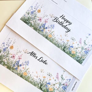 K&ouml;nnte beinhalten: Zwei wei&szlig;e Gru&szlig;karten mit floralen Aquarelldesigns. Eine Karte tr&auml;gt den Schriftzug "Happy Birthday" in schwarzer Schrift. Die andere Karte tr&auml;gt den Schriftzug "Alles Liebe". Die Karten zeigen bunte Blumen und Schmetterlinge.