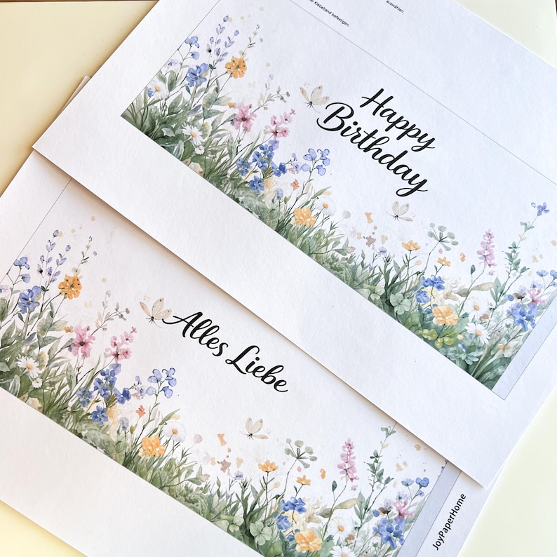 K&ouml;nnte beinhalten: Zwei wei&szlig;e Gru&szlig;karten mit floralen Aquarelldesigns. Eine Karte tr&auml;gt den Schriftzug "Happy Birthday" in schwarzer Schrift. Die andere Karte tr&auml;gt den Schriftzug "Alles Liebe". Die Karten zeigen bunte Blumen und Schmetterlinge.