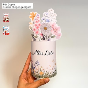 K&ouml;nnte beinhalten: Ein wei&szlig;er zylindrischer Beh&auml;lter mit floralem Design und dem Schriftzug "Alles Liebe" in schwarzer Schrift. Der Beh&auml;lter enth&auml;lt Papierblumen in Rosa-, Gelb- und Blaut&ouml;nen. Der Text "F&uuml;r Duplo Kinder Riegel geeignet" befindet sich oben links.