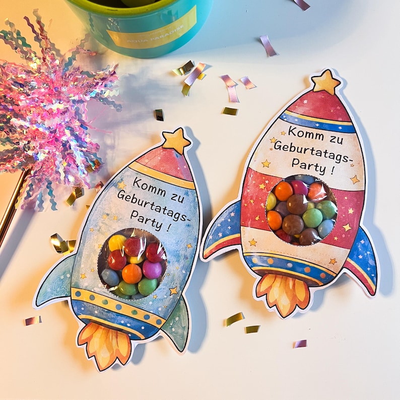 May include: Two colorful rocket-shaped party favor bags filled with candies. The rockets are red, blue, and yellow with the text "Komm zu Geburtstags-Party!" printed on them. Confetti is scattered around the rockets.