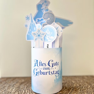 Könnte beinhalten: Zylindrische Geburtstagsdekoration mit hellblauem Aquarelldesign und dem Schriftzug "Alles Gute zum Geburtstag" in blauer Schrift. Mit Ausschnitten von Schneeflocken, einem Schloss und der Zahl 2.