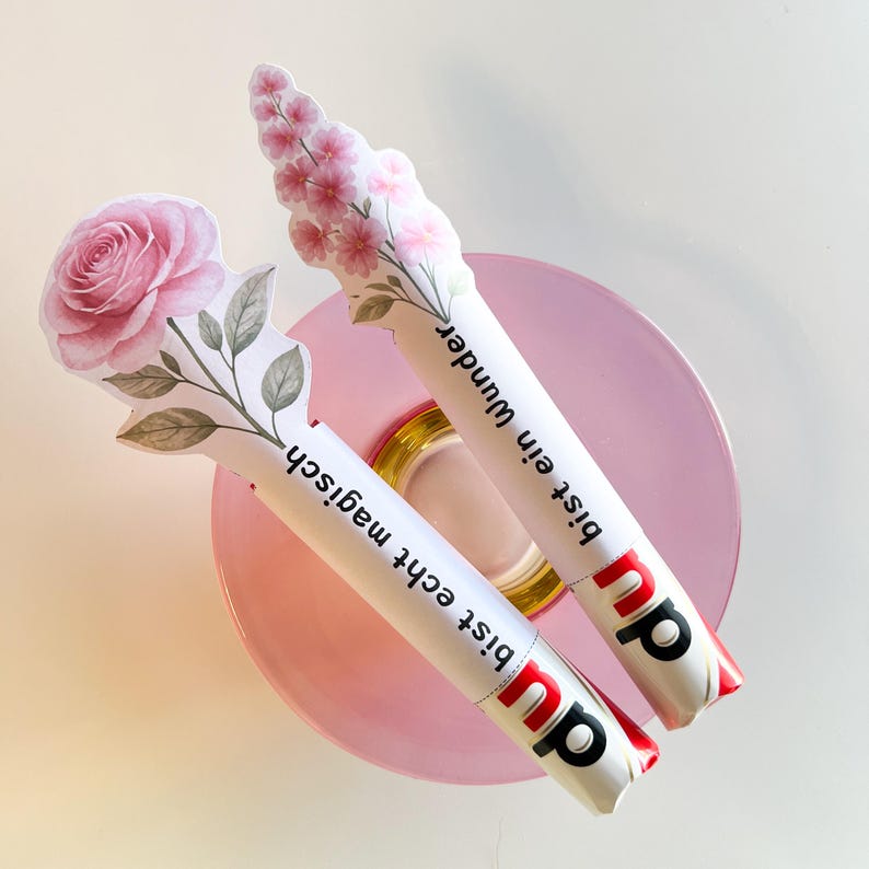 K&ouml;nnte beinhalten: Zwei wei&szlig;e Schokoriegel mit floralen Designs und deutschem Text. Einer zeigt eine rosa Rose, der andere rosa Bl&uuml;ten. Die Riegel liegen auf einem rosa Teller, mit dem Markenlogo sichtbar.