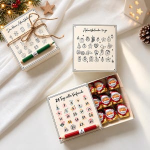 Könnte beinhalten: Drei rechteckige Adventskalender-Schachteln mit cremefarbenen Deckeln. Zwei Schachteln zeigen schwarze Strichzeichnungen von Weihnachtsmotiven mit den Zahlen 1 bis 24 und einem grünen oder roten Stift. Eine offene Schachtel enthält neun Schoko-Weihnachtsmänner.