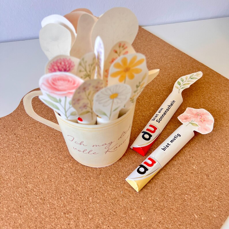 May include: A paper flower arrangement in a cream-colored cup with the text "Ich mag die volle Kanne". Two paper bookmarks with the text "du bist ein Sonnenschein" and "du bist mutig" are also present. The flowers include roses and daisies.