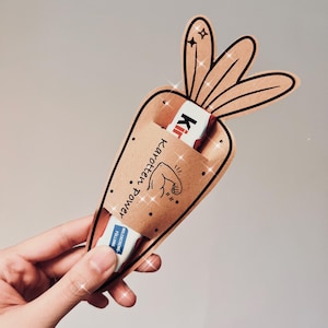 May include: A brown paper carrot-shaped holder with a bunny ear design, containing two Kinder chocolate bars. The holder features the text "Karotten Power" and an illustration of a flexing arm. The design has black outlines and sparkles.