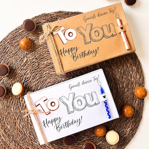 Peut inclure: Deux boîtes cadeaux rectangulaires avec le texte To You et Happy Birthday, accompagnées d'une bougie chacune. Une boîte est en carton brun avec une bougie rose, l'autre est blanche avec une bougie bleue à pois, entourées de chocolats assortis.