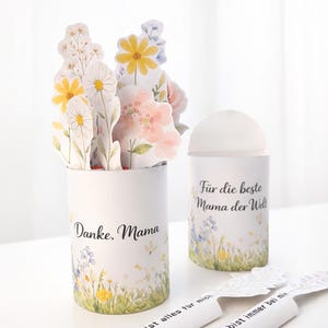 May include: Two white cylindrical containers with floral designs and text. One says "Danke, Mama" and holds paper flowers. The other says "Für die beste Mama der Welt". Additional paper strips with text are also visible.