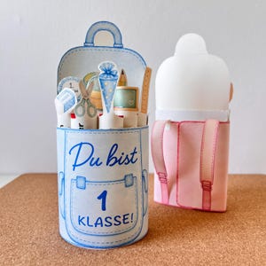 May include: A light blue and white paper pencil holder shaped like a backpack, with the words "Du bist 1 Klasse!" in blue script. The holder contains paper cutouts of school supplies. A pink backpack is next to it.