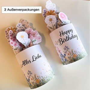 K&ouml;nnte beinhalten: Zwei wei&szlig;e zylindrische Geschenkboxen mit Blumendesigns und Pop-up-Blumenarrangements. Eine Box tr&auml;gt die Aufschrift "Happy Birthday" und die andere "Alles Liebe". Die Boxen sind mit Blumen im Aquarellstil verziert.