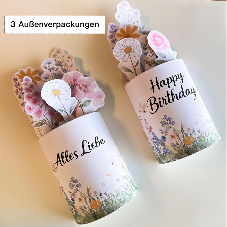 K&ouml;nnte beinhalten: Zwei wei&szlig;e zylindrische Geschenkboxen mit Blumendesigns und Pop-up-Blumenarrangements. Eine Box tr&auml;gt die Aufschrift "Happy Birthday" und die andere "Alles Liebe". Die Boxen sind mit Blumen im Aquarellstil verziert.
