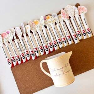 May include: A collection of decorative spoon-shaped bookmarks with floral designs and German text, arranged on a brown corkboard. A small, cream-colored pitcher with the text "Ich mag dich volle Kanne" is also present.
