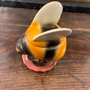 May include: A ceramic bee-shaped trinket box, featuring a white, orange, and black body. The lid is white and curved, mimicking wings. The box rests on a small, scalloped, pink base. This bee-shaped box is a decorative item.