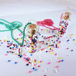 May include: Two glass bottles filled with colorful sprinkles, each attached to a chain necklace. One necklace is teal and the other is pink. The bottles are on a white surface with scattered sprinkles.