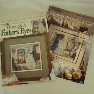 May include: Two cross-stitch pattern leaflets from Leisure Arts. One is titled "Through A Father's Eyes" and the other "Through A Mother's Eyes." Both feature framed cross-stitch designs with a wedding theme. The leaflets are on a white surface.