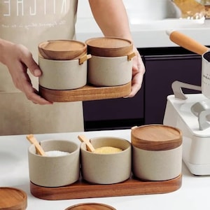 Könnte beinhalten: Ein Set aus beigen Keramik-Gewürzgläsern mit Holzdeckeln und kleinen Holzspateln. Die Gläser stehen auf einem Holztablett. Das Bild zeigt zwei Sets, eines mit zwei Gläsern und das andere mit drei.